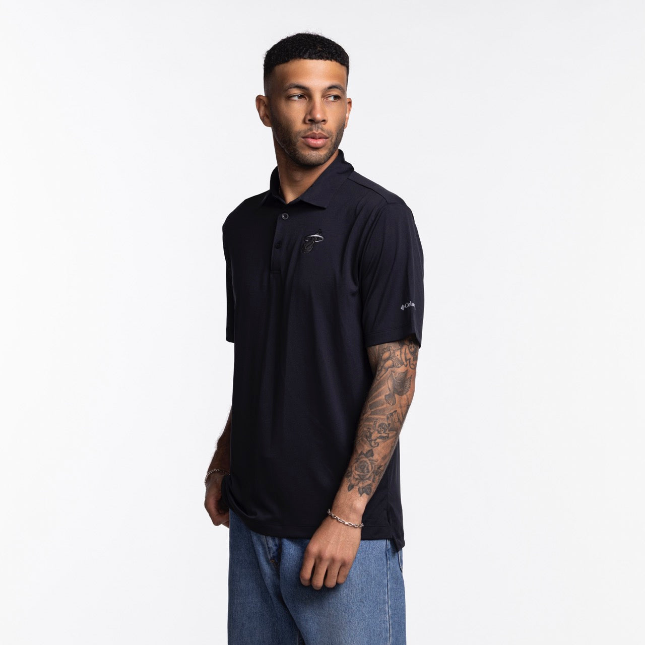 A man with short dark hair and a tattooed right arm wears the Columbia Outdoor Miami HEAT Original Vice Nights Tonal Polo in black, paired with blue jeans, standing against a plain white background and looking slightly left.