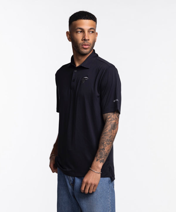 A man with short hair, trimmed beard, and tattooed forearm wears the Columbia Outdoor Miami HEAT Original Vice Nights Tonal Polo—black, standard fit, short-sleeve—paired with light blue jeans against a plain white background.