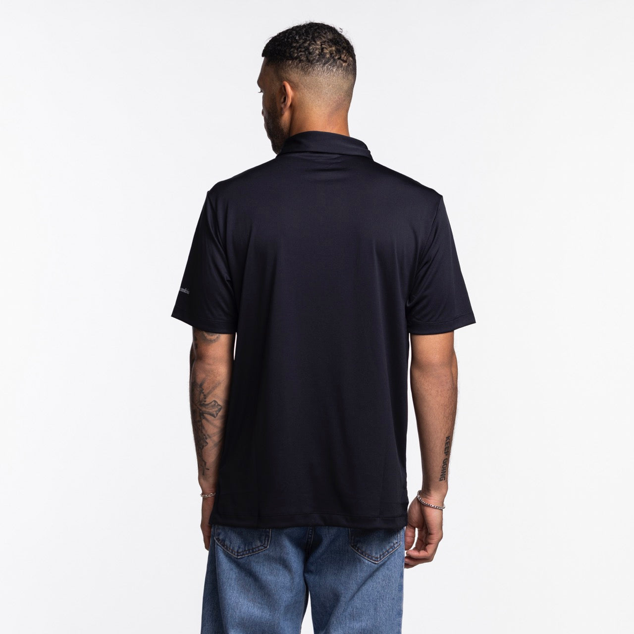 A man with short hair and arm tattoos faces away from the camera, wearing a Columbia Outdoor Miami HEAT Original Vice Nights Tonal Polo in black and blue jeans, against a plain white background.