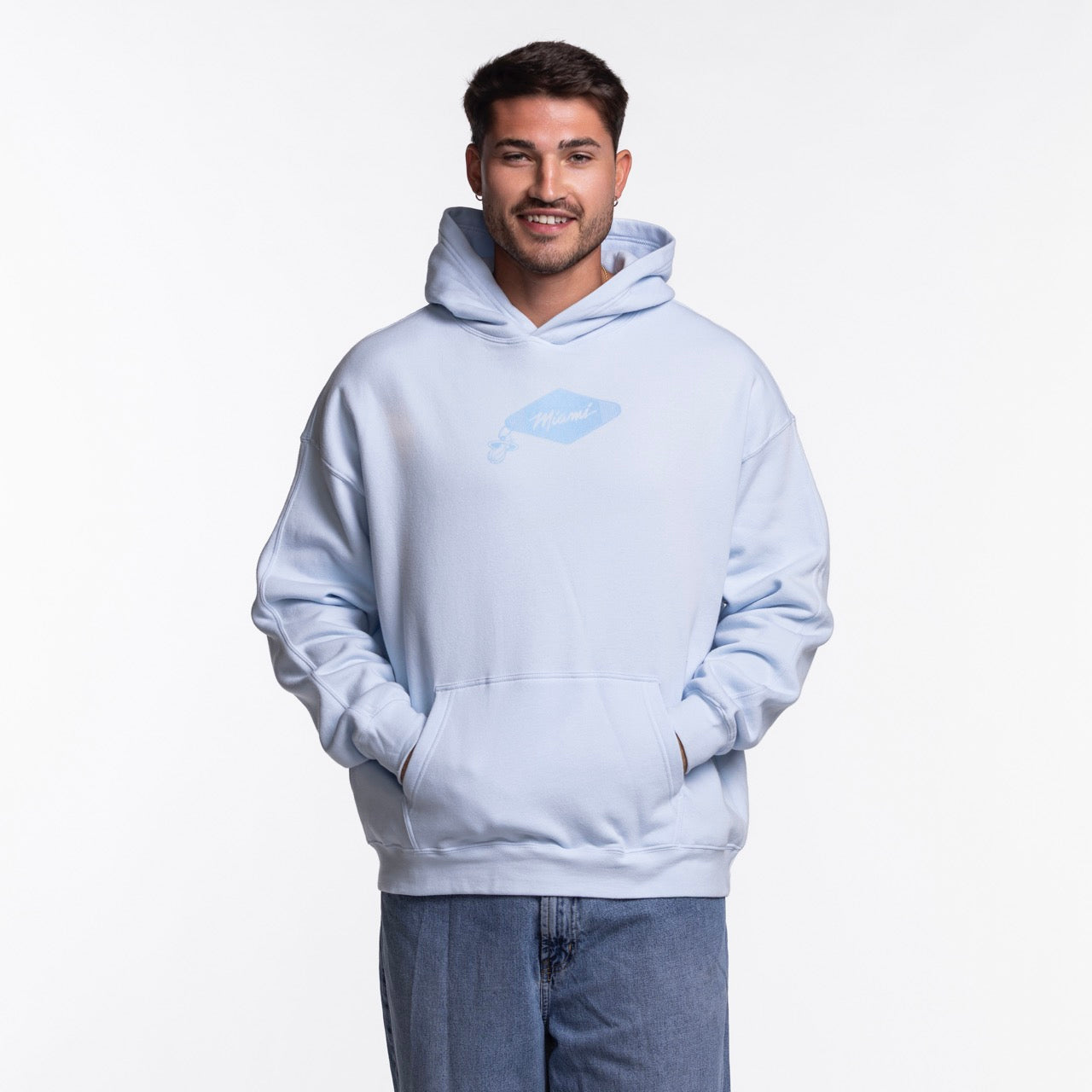 A man stands facing forward, smiling with hands in the pocket of a light blue Court Culture Original Vice Nights 