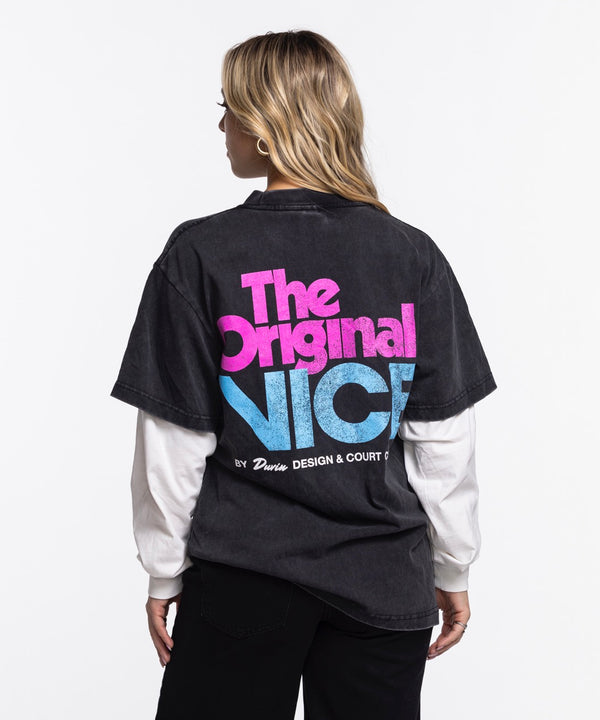 A man stands against a white background wearing a black Court Culture x Duvin Original Vice Long Sleeve Tee by Court Culture, featuring pink Vice Nights lettering, layered over a white long-sleeve shirt with black pants.