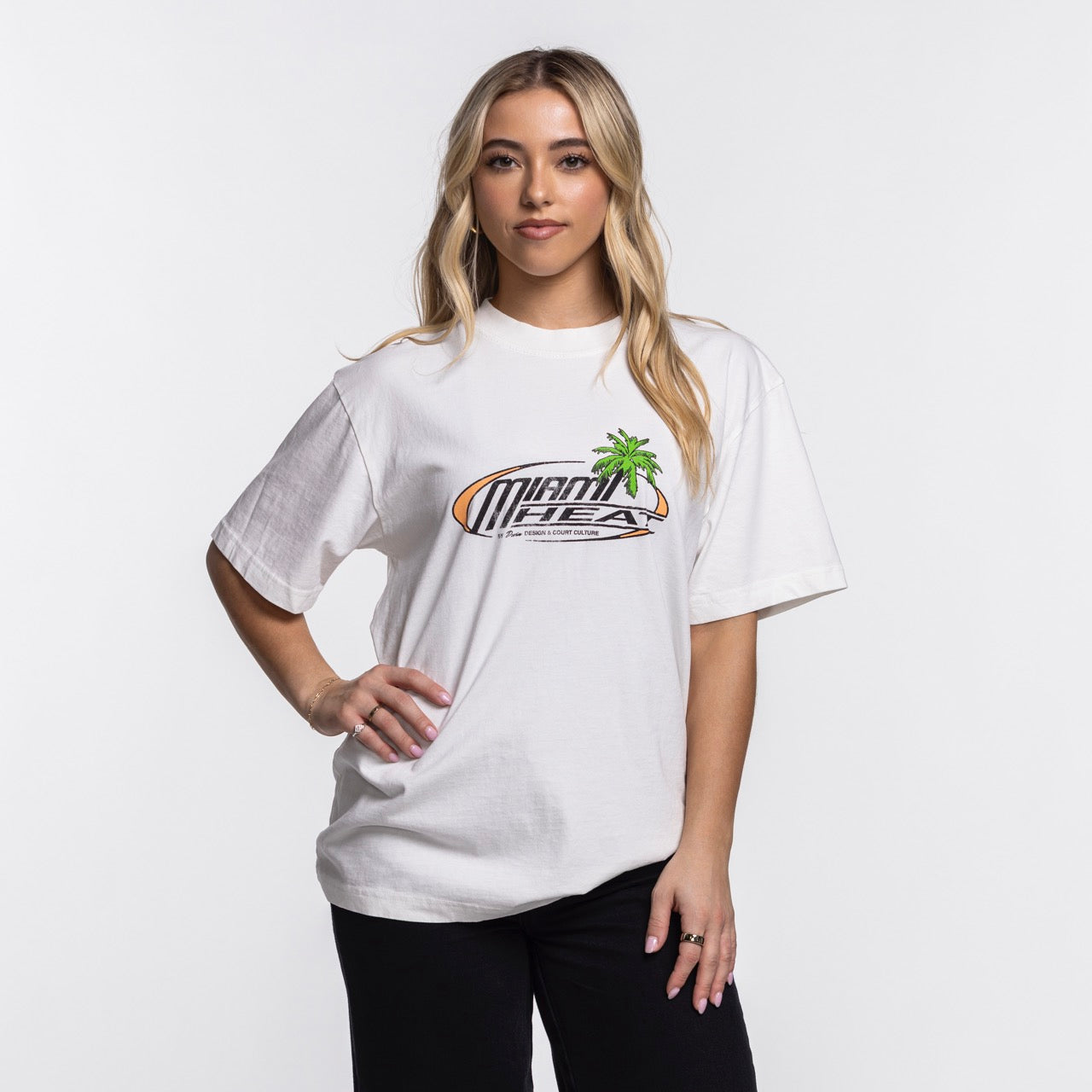 A woman with long blonde hair wears the white Court Culture x Duvin Sun Tee by Court Culture, paired with black pants, standing against a plain white background with one hand on her hip.