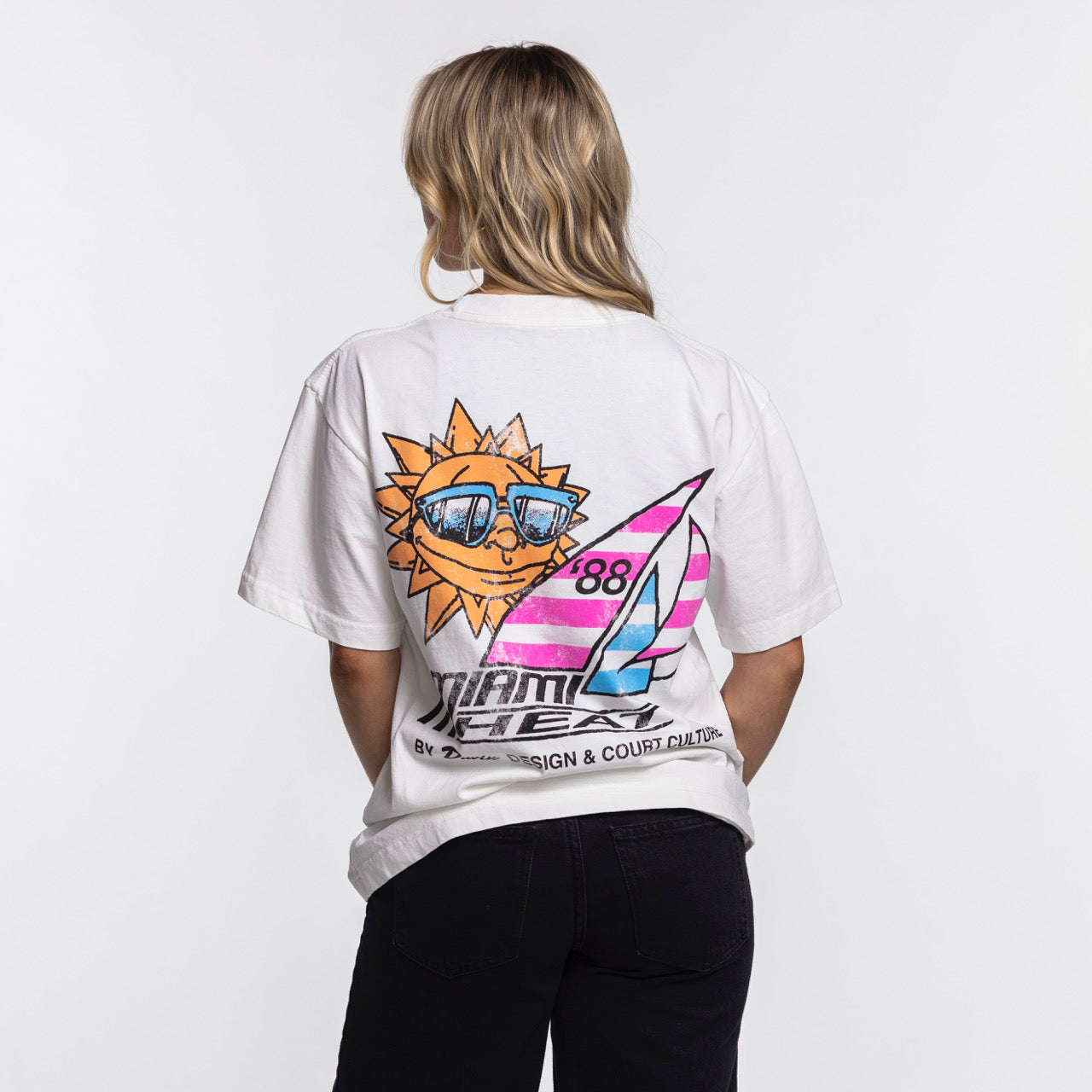 A person with blonde hair, seen from behind, wears the Court Culture x Duvin Sun Tee by Court Culture, featuring a colorful graphic of a smiling sun with sunglasses, sailboat, number 88, and unique stylized text and designs.