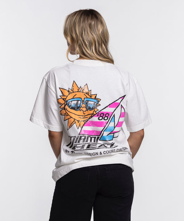 A man stands against a plain white background, wearing the Court Culture x Duvin Sun Tee from Court Culture with a Miami Heat logo and palm tree graphic, paired with black pants. He looks to the side and smiles.