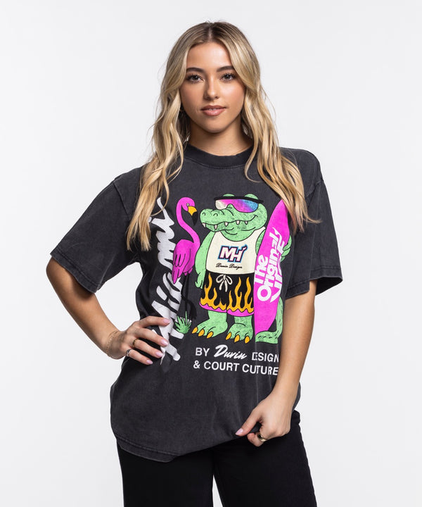 A man stands against a white background wearing the Court Culture x Duvin Flame Surf Tee by Court Culture, a black heavyweight shirt featuring colorful graphics of a cartoon crocodile, flamingo, surfboard, and 