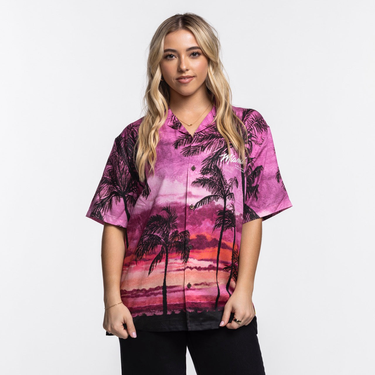 A woman with long blonde hair wears the Court Culture x Duvin Sunset Button-Up by Court Culture, featuring vibrant pink and purple hues, as she stands against a plain white background, looking at the camera with a slight smile.
