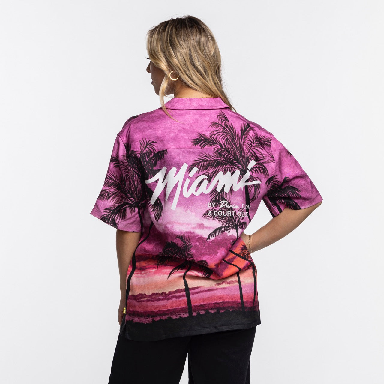 A blonde-haired person stands facing away, wearing the Court Culture x Duvin Sunset Button-Up from Court Culture, featuring palm trees, bold 