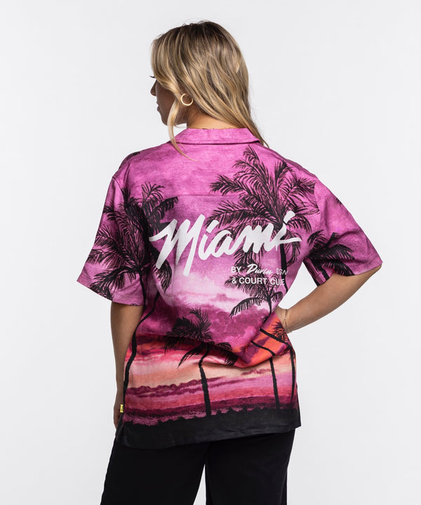 A man wears the Court Culture x Duvin Sunset Button-Up, featuring a vibrant pink and purple sunset with palm tree design, standing against a white background and looking left with one hand in his pocket.