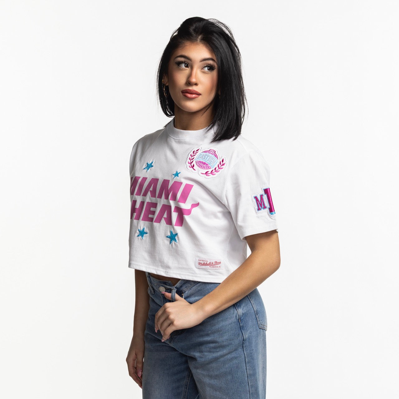 A woman with straight black hair wears a Mitchell & Ness Miami HEAT Original Vice Nights Women's Boxy Tee and blue jeans, standing against a plain white background.