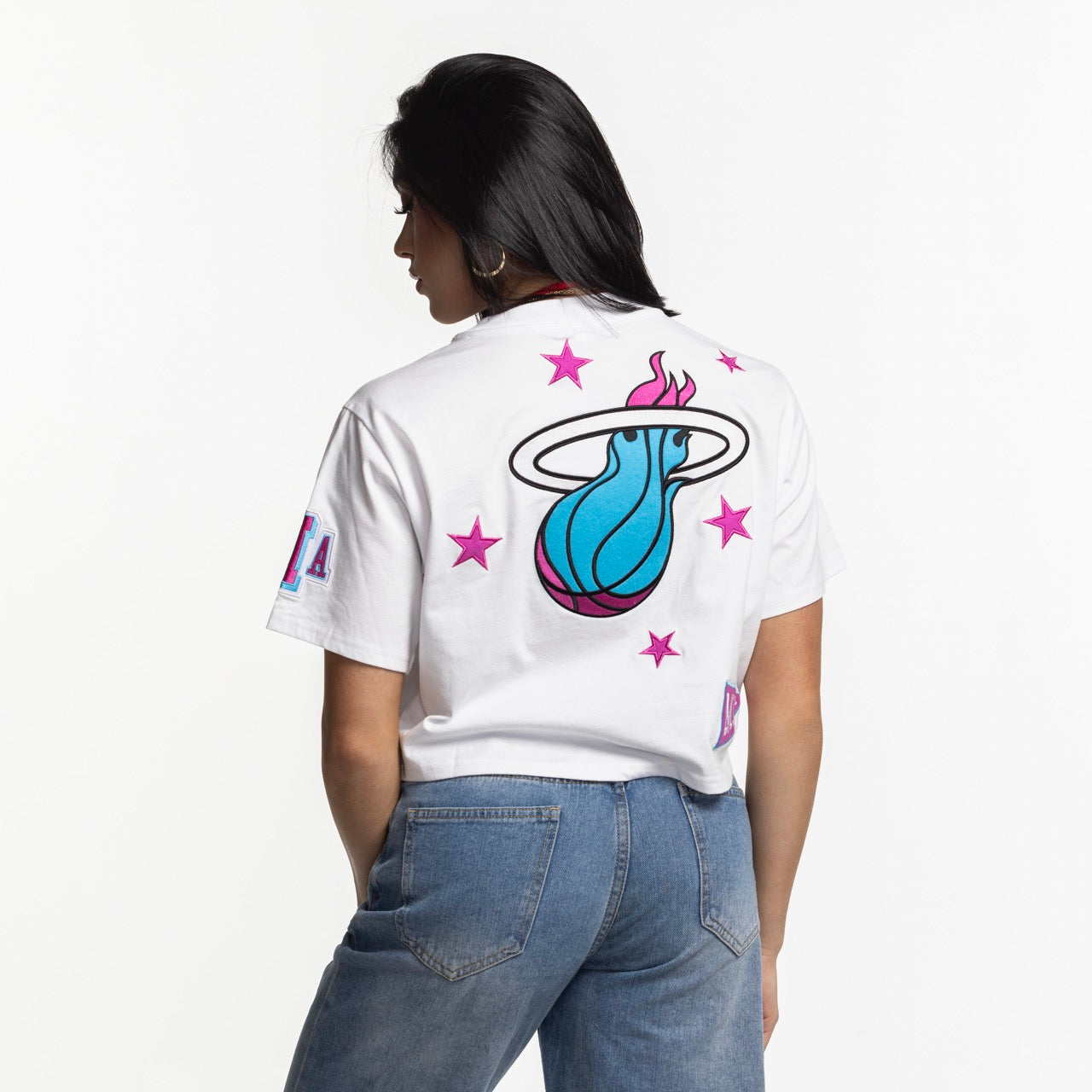 A woman with long dark hair wears the Mitchell & Ness Miami HEAT Original Vice Nights Women's Boxy Tee—a white, boxy cotton T-shirt with a large blue and pink flaming basketball logo and stars on the back—paired with blue jeans against a white background.