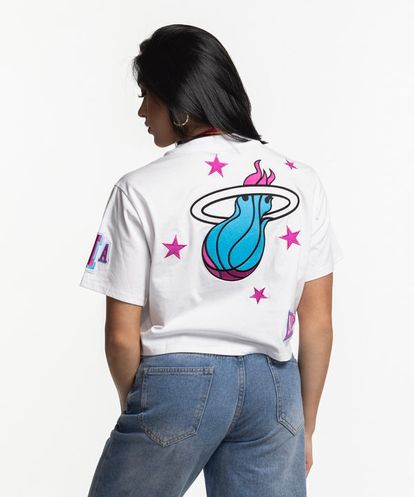 A woman with straight black hair and hoop earrings smiles in blue jeans and a Mitchell & Ness Miami HEAT Original Vice Nights Women's Boxy Tee against a plain white background.