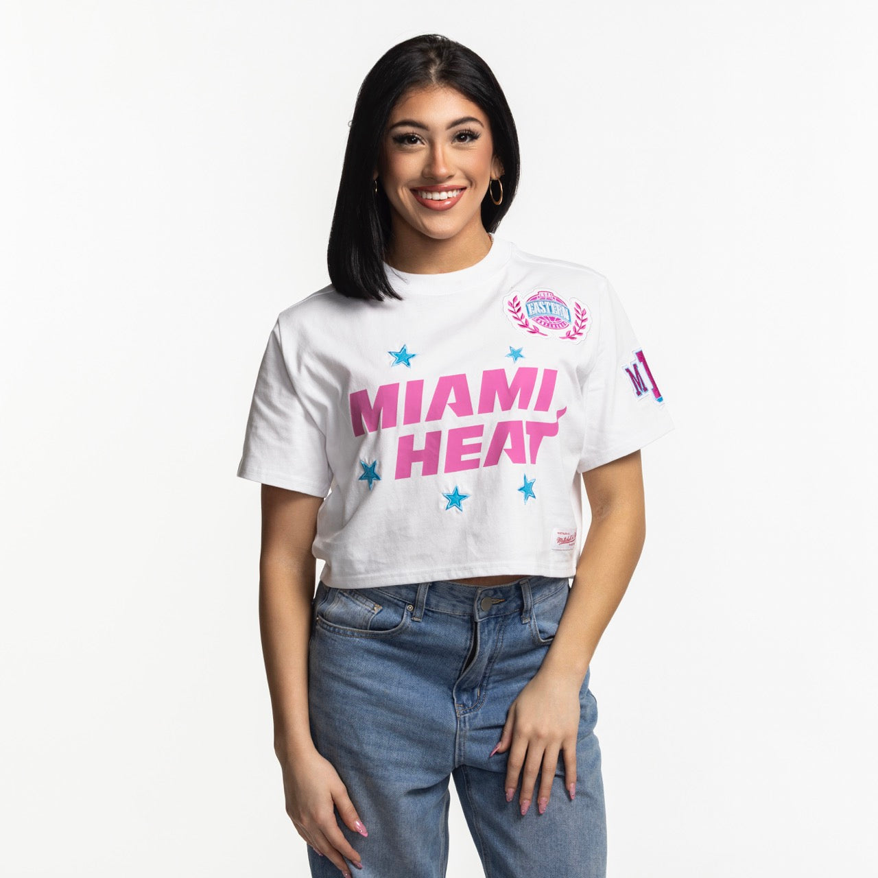 A woman with straight black hair and hoop earrings smiles in blue jeans and a Mitchell & Ness Miami HEAT Original Vice Nights Women's Boxy Tee against a plain white background.
