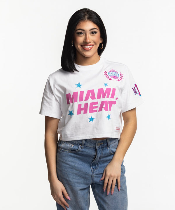 A woman with straight black hair and hoop earrings smiles in blue jeans and a Mitchell & Ness Miami HEAT Original Vice Nights Women's Boxy Tee against a plain white background.