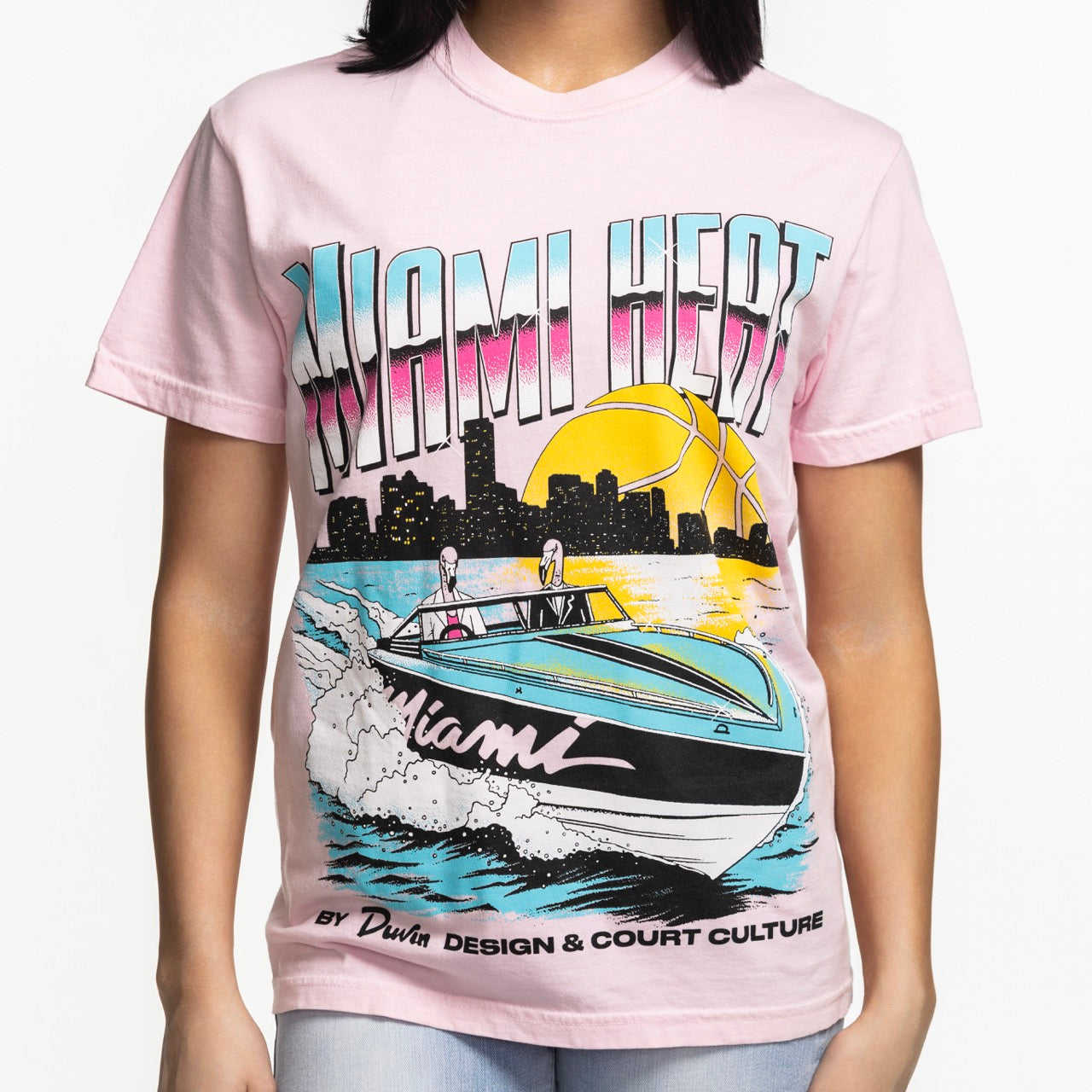 A person wears the Court Culture x Duvin Boat Tee by Court Culture, a pink tee with a vibrant speedboat scene, city skyline, yellow basketball, bold retro text—all crafted from soft, pre-shrunk midweight cotton for lasting comfort.