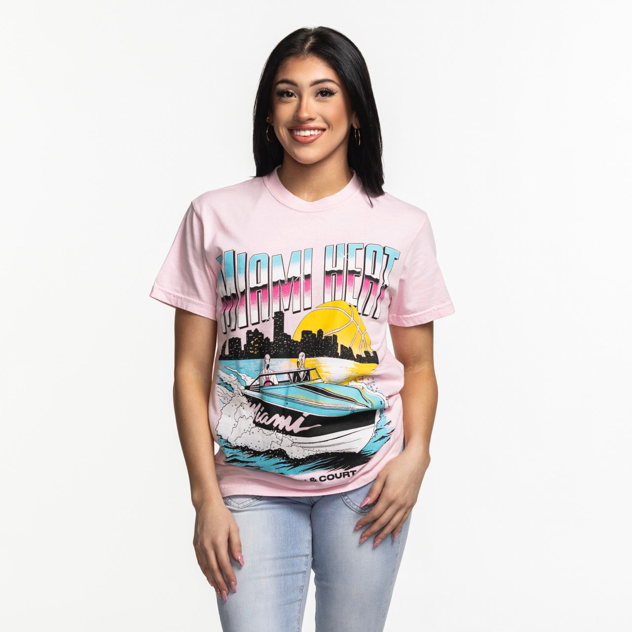 A woman with long dark hair wears the Court Culture x Duvin Boat Tee by Court Culture, featuring a colorful boat, sunset, and city skyline graphic. She pairs it with light blue jeans and stands against a plain white background.
