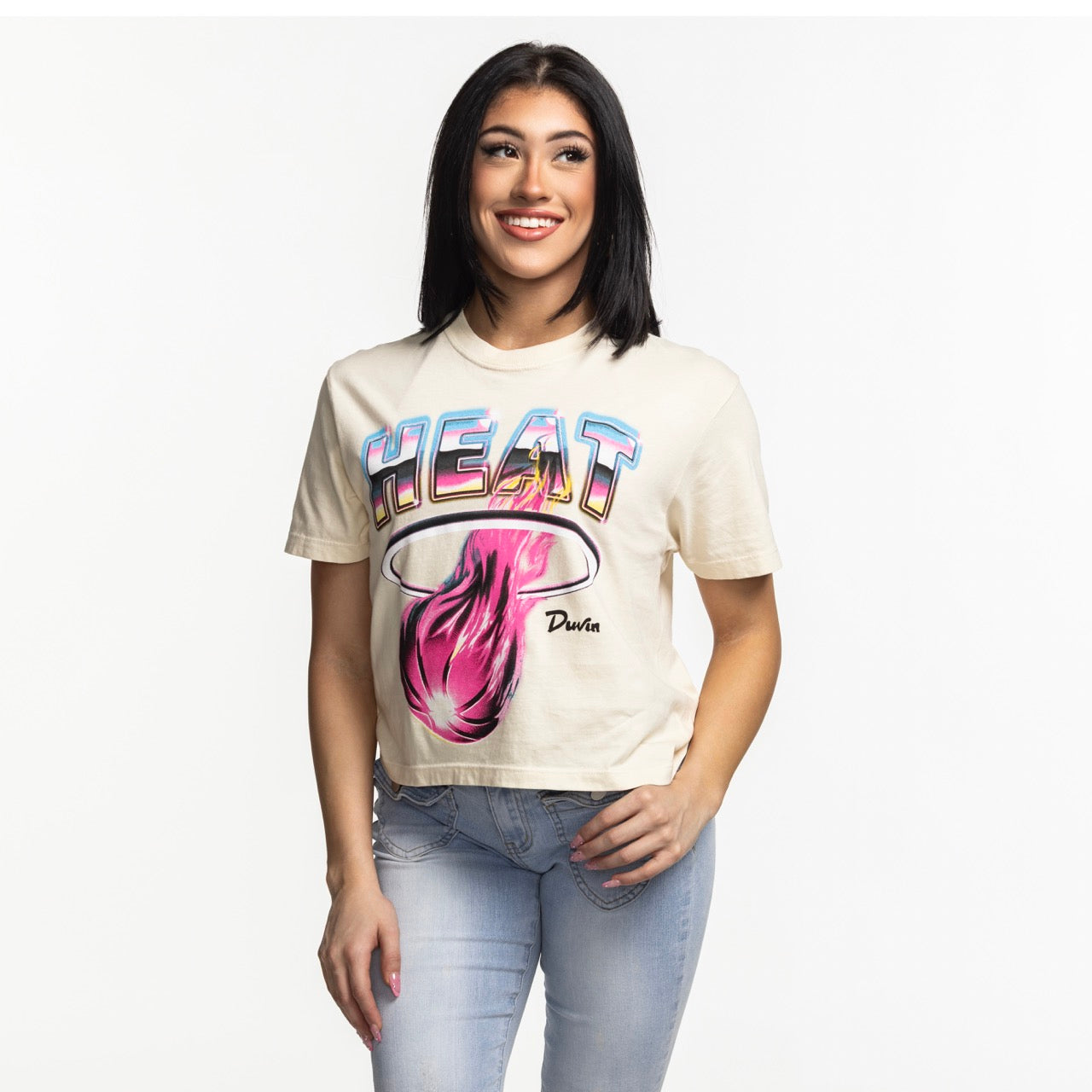 A woman with straight dark hair, smiling with one hand in her jeans pocket, wears the beige Court Culture x Duvin Tropical Crop tee against a plain white background.