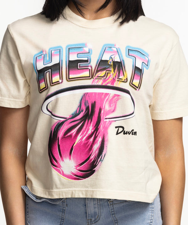 A woman with straight dark hair, smiling with one hand in her jeans pocket, wears the beige Court Culture x Duvin Tropical Crop tee against a plain white background.