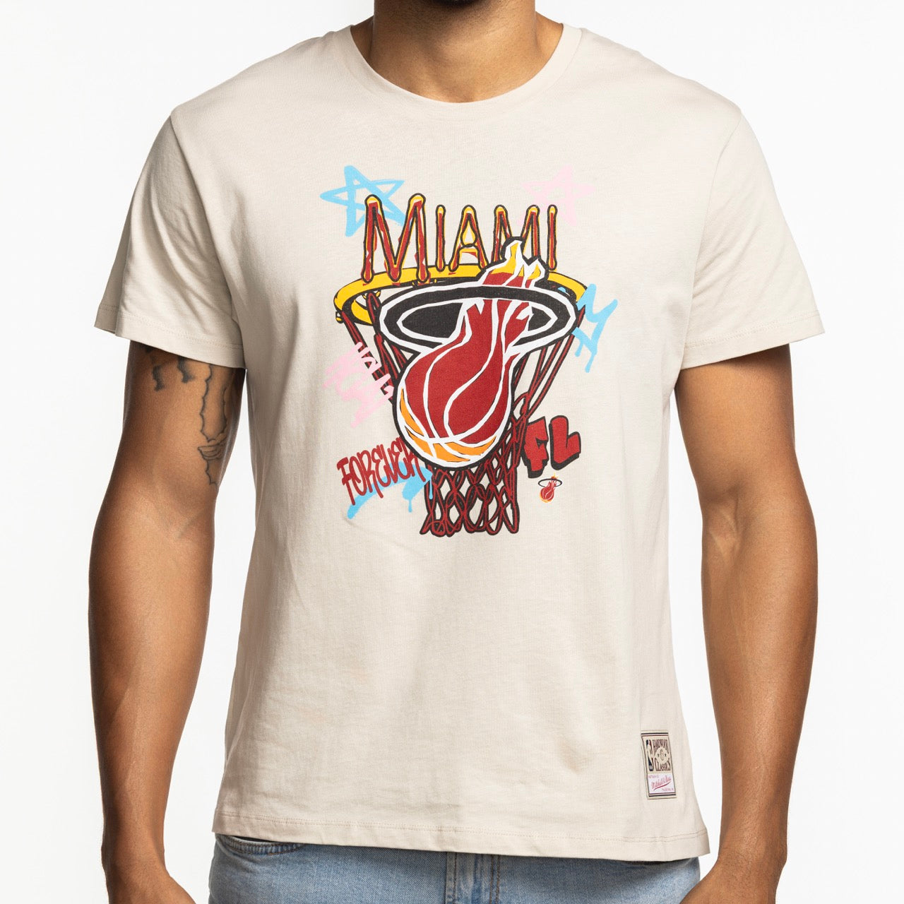 A person wearing the Mitchell & Ness Miami HEAT City Dreams Boxy Tee—a beige, standard fit cotton T-shirt with a colorful flaming basketball graphic, MIAMI text, and blue/pink graffiti accents. Machine washable for easy care.