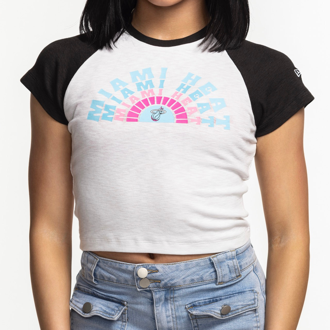 A woman wears the New Era Miami HEAT Original Vice Women's Raglan Tee—a snug, cropped white and black t-shirt with pastel text and logo—paired with high-waisted blue jeans. Made from a cotton-modal-spandex blend. Background is plain white.