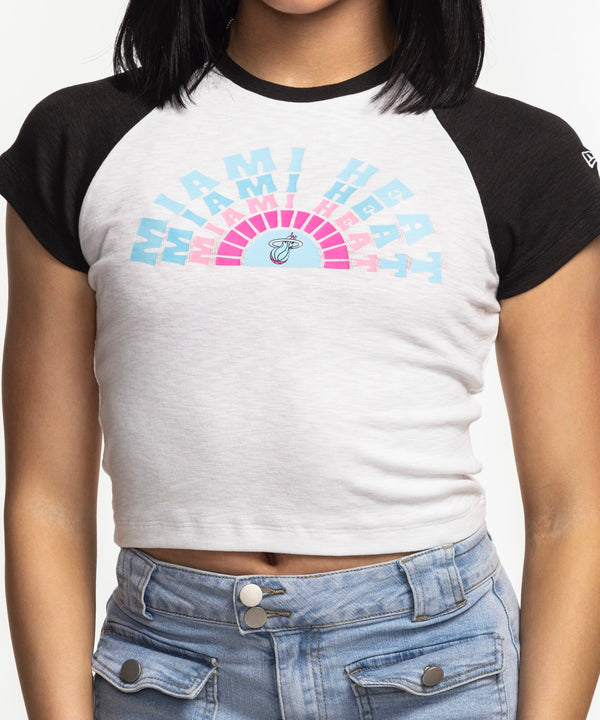 A woman with straight black hair wears the New Era Miami HEAT Original Vice Women's Raglan Tee and light blue jeans, standing against a plain white background, gazing slightly to the side with one hand on her hip.
