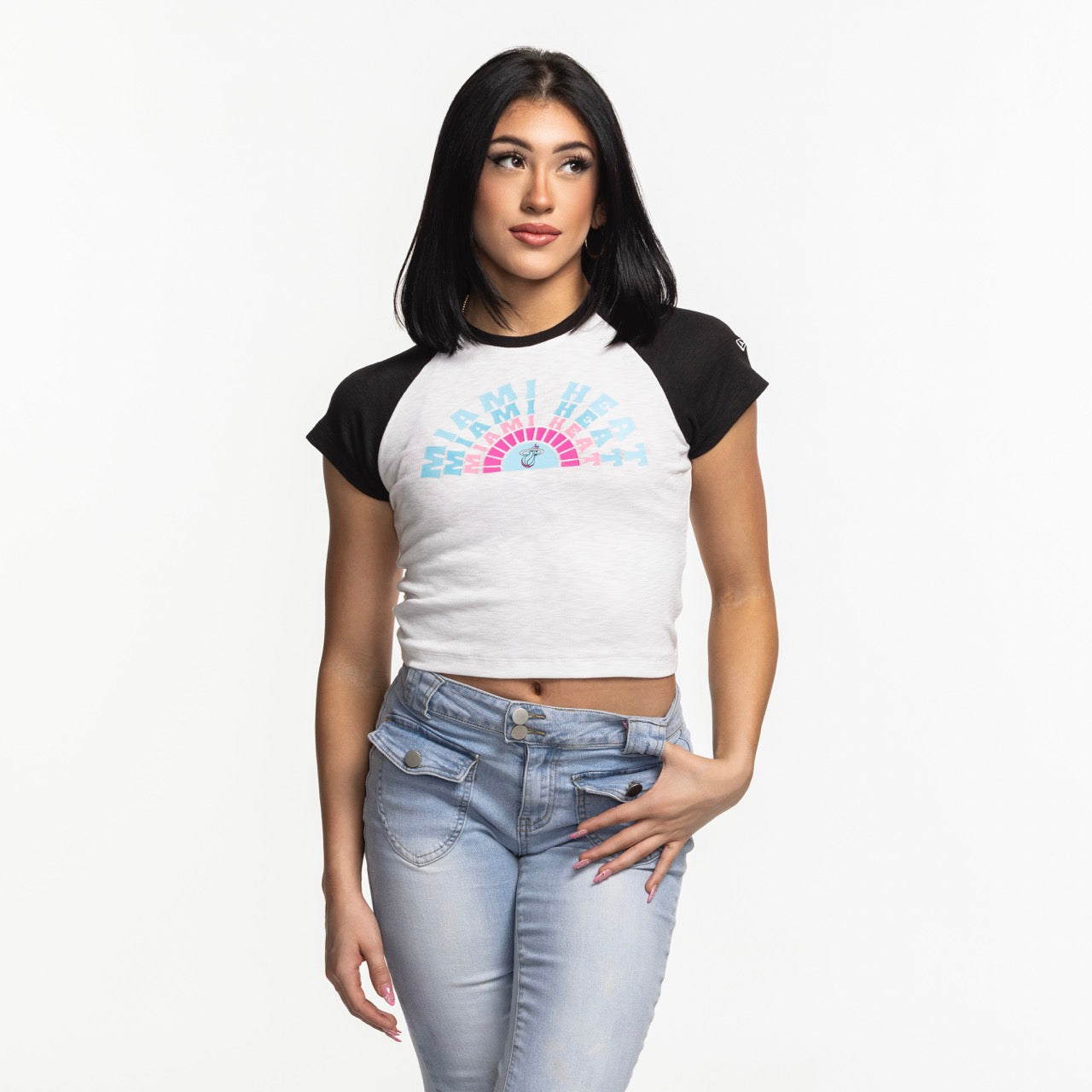 A woman with straight black hair wears the New Era Miami HEAT Original Vice Women's Raglan Tee and light blue jeans, standing against a plain white background, gazing slightly to the side with one hand on her hip.