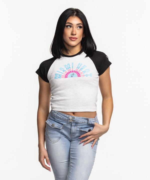 A woman with straight black hair wears the New Era Miami HEAT Original Vice Women's Raglan Tee and light blue jeans, standing against a plain white background, gazing slightly to the side with one hand on her hip.