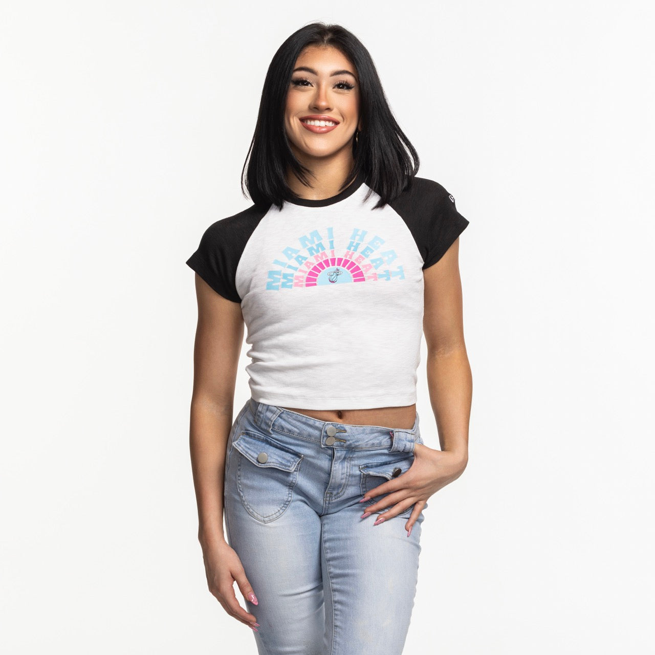 A woman with straight black hair smiles while wearing a snug-fitting New Era Miami HEAT Original Vice Women's Raglan Tee and light blue jeans, standing against a plain white background.