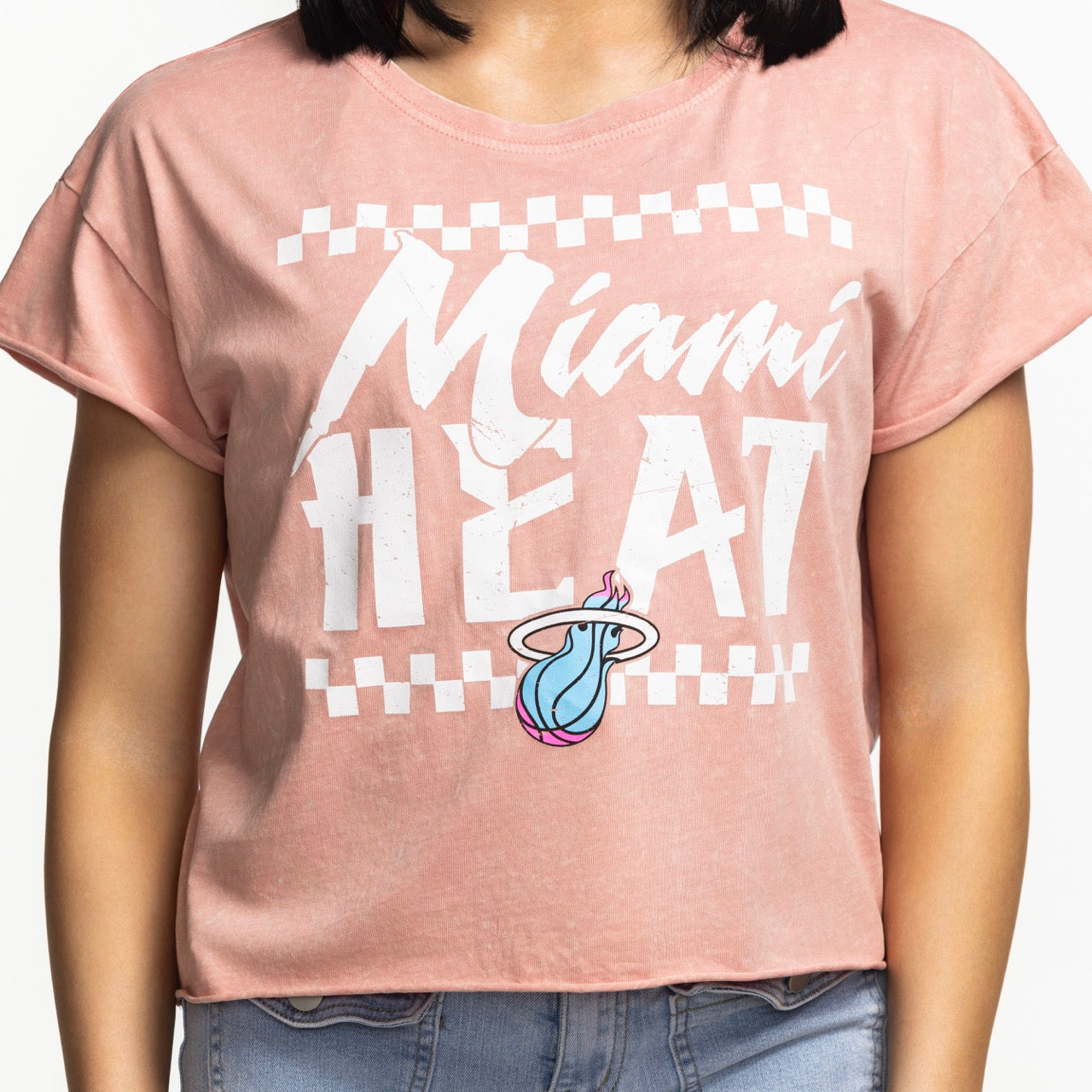 A person wears the Sportiqe Miami HEAT Original Vice Coral Women's Tee—a light pink, relaxed-fit T-shirt by Sportiqe with a flaming basketball logo—paired with light blue jeans. Made of cotton, it's machine washable. The person's head is not visible.