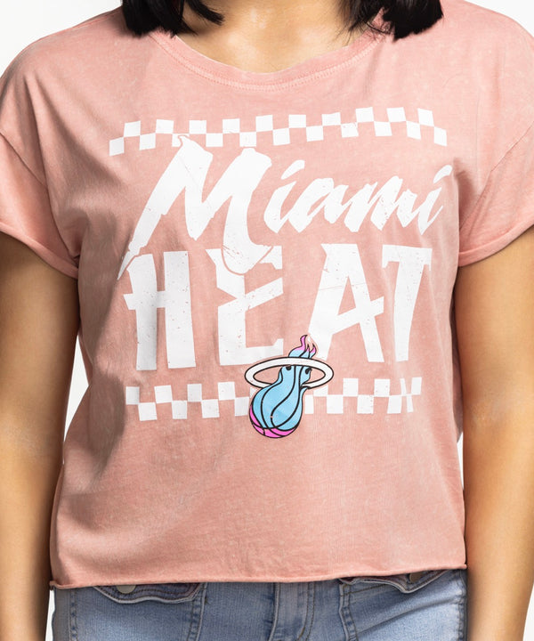 A woman with straight dark hair wears the Sportiqe Miami HEAT Original Vice Coral Women's Tee by Sportiqe, paired with light blue jeans. Standing against a white background with one hand in her pocket. Easy care—just machine wash.