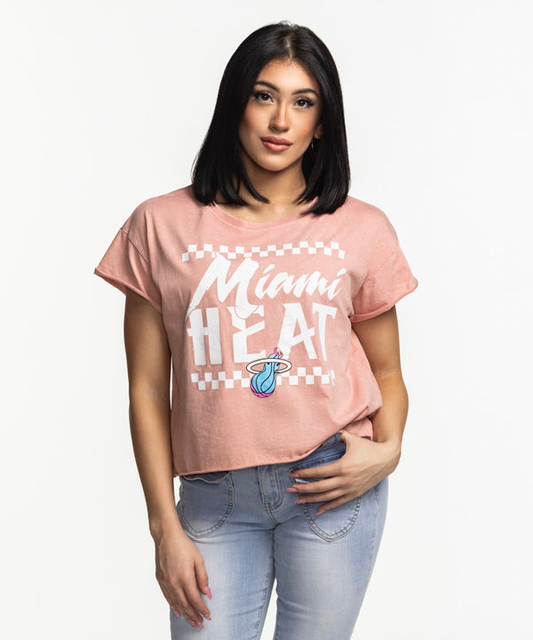 A woman with straight dark hair wears the Sportiqe Miami HEAT Original Vice Coral Women's Tee by Sportiqe, paired with light blue jeans. Standing against a white background with one hand in her pocket. Easy care—just machine wash.