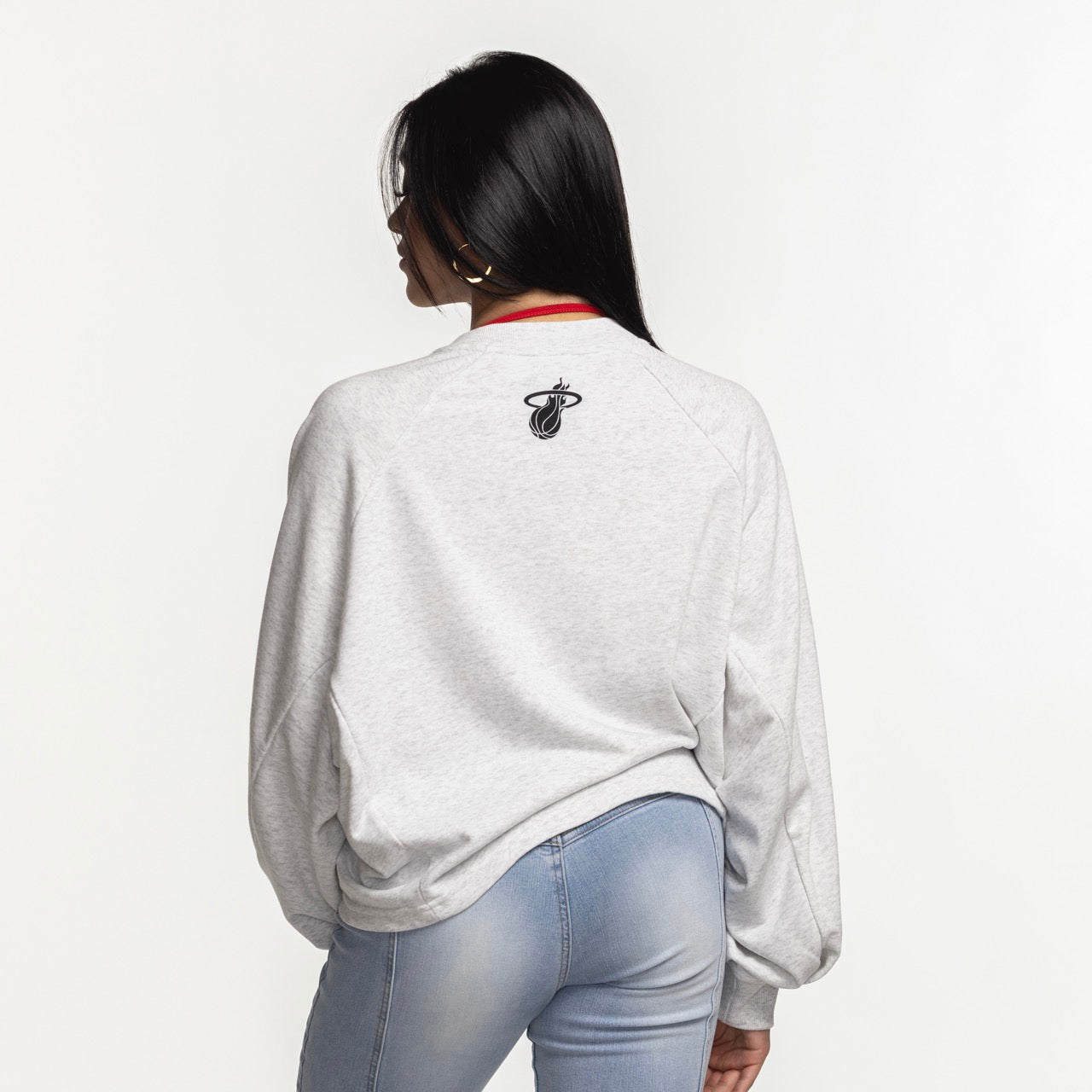 A woman with long dark hair wears the G-III Miami HEAT Faceoff Women's Crewneck by G-III Sports—a relaxed fit light gray cotton-polyester sweatshirt with a small basketball logo on the upper back—paired with light blue jeans, facing away on a white background.