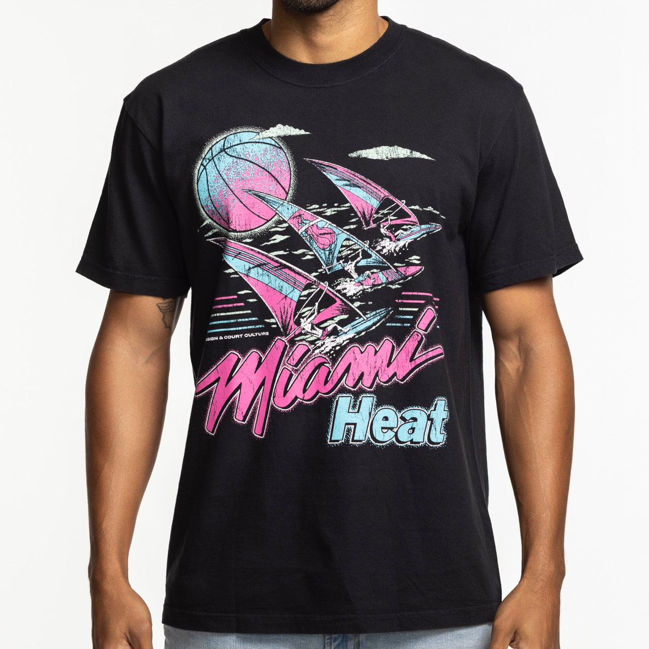 Someone wearing the Court Culture x Duvin Tropical Tee by Court Culture—a black mid-weight cotton shirt with a retro Miami Heat graphic featuring pink, blue, and white sailboats, a basketball, and stylized waves.
