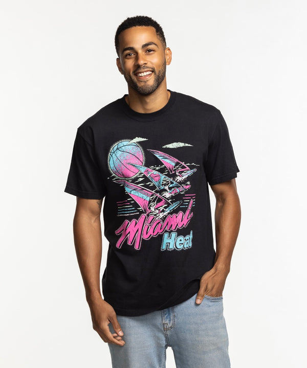 A man smiles in the Court Culture x Duvin Tropical Tee by Court Culture, a black mid-weight cotton shirt with a retro basketball, palm trees, and Miami Heat text, paired with light blue jeans against a plain white background.