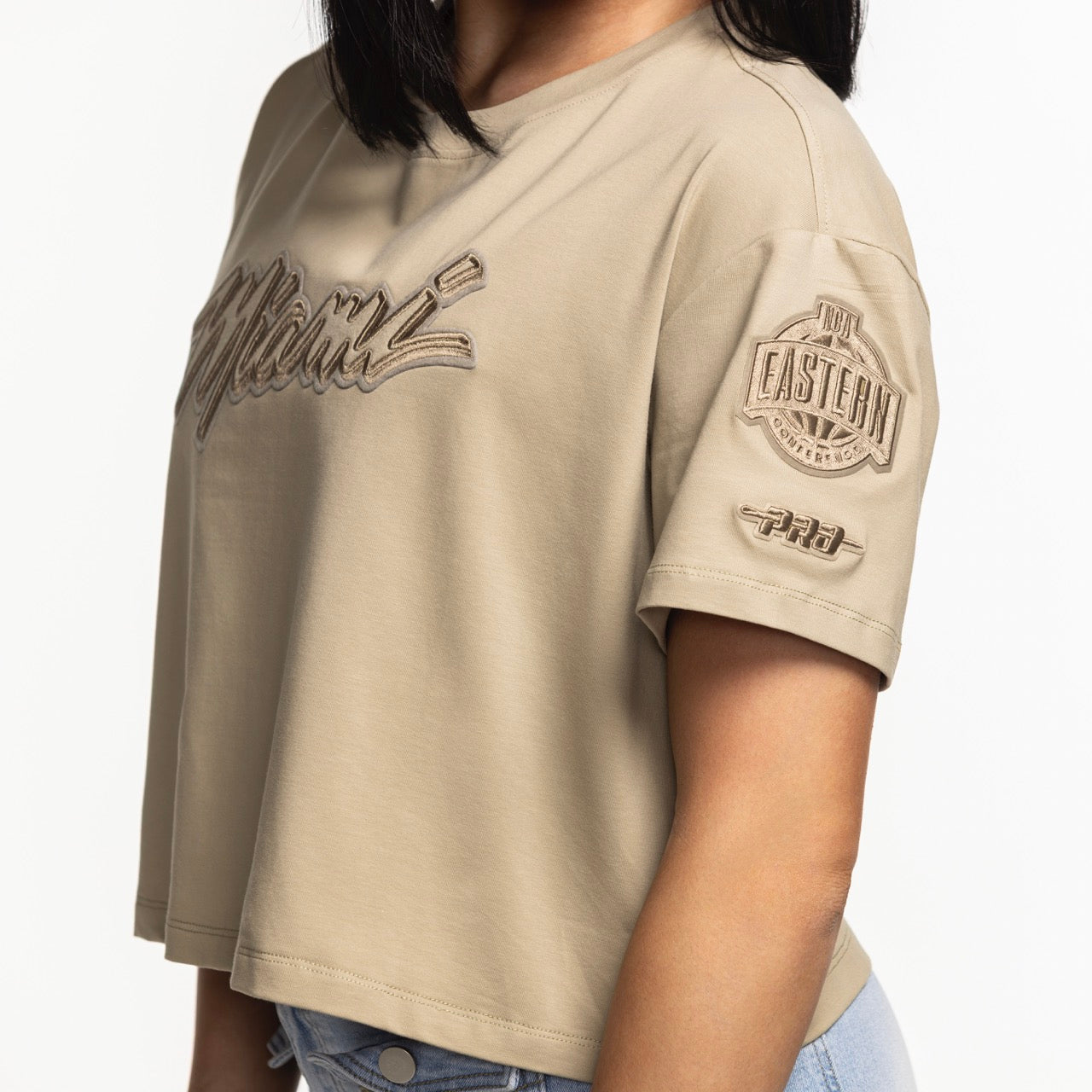 A woman wearing the Pro Standard Miami HEAT Original Vice Nights Beige Women's Boxy Tee, a cropped cotton top with embroidered text, and light blue jeans, is shown from shoulders to waist against a white background.