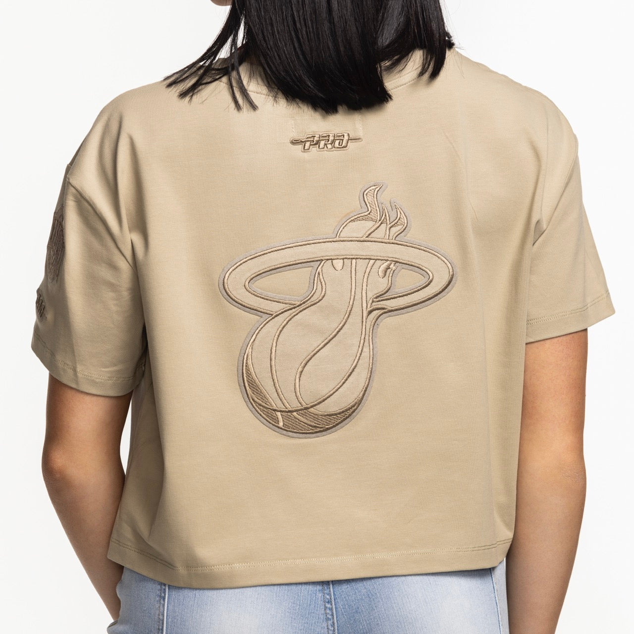 A woman wears the Pro Standard Miami HEAT Original Vice Nights Beige Women's Boxy Tee, featuring the team logo on the back, styled with light blue jeans. Photo is taken from behind to showcase its boxy cropped fit. Brand: Pro Standard.