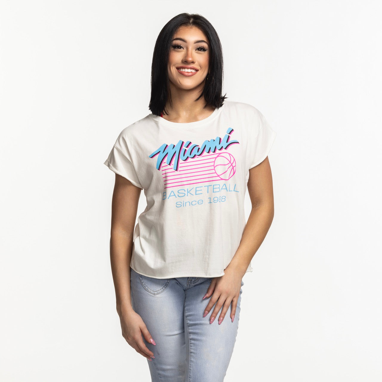 A woman with straight dark hair smiles, wearing a Sportiqe Miami HEAT Original Vice Nights Alana Women's Tee and light blue ripped jeans, standing against a plain white background.