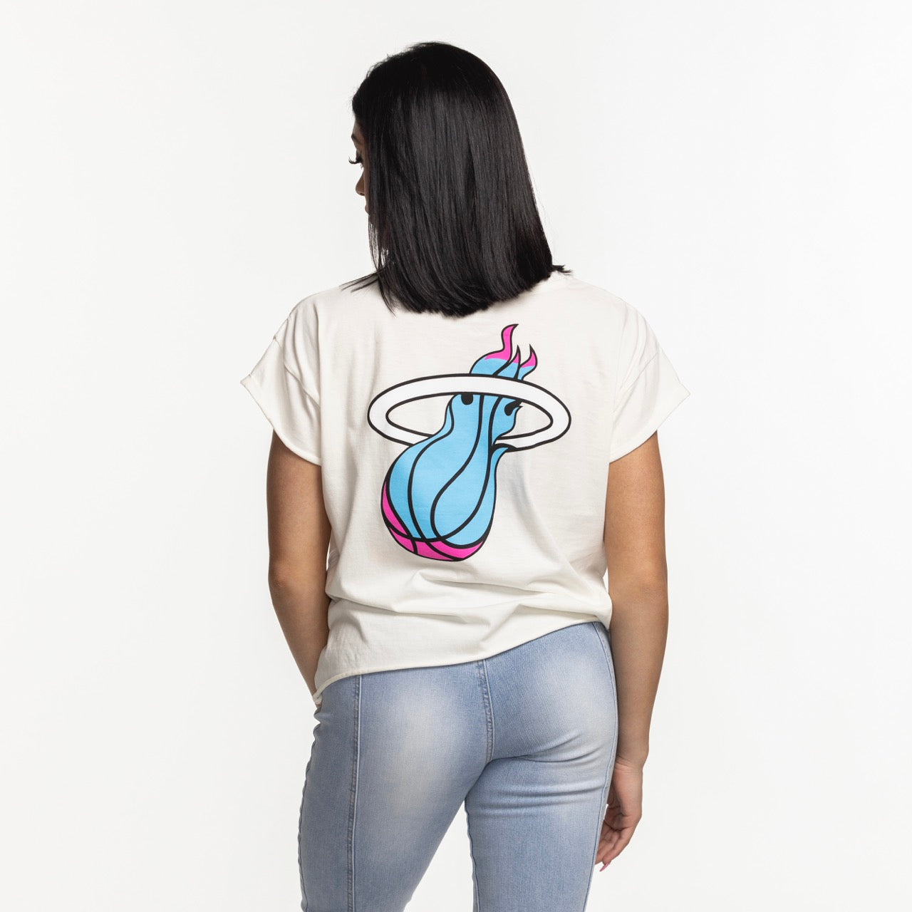 A woman with straight black hair faces away, wearing the Sportiqe Miami HEAT Original Vice Nights Alana Women's Tee featuring a blue, pink, and black flaming basketball logo on the back, paired with light blue jeans against a white background.