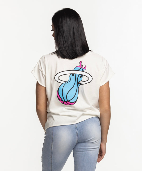 A woman with straight dark hair smiles, wearing a Sportiqe Miami HEAT Original Vice Nights Alana Women's Tee and light blue ripped jeans, standing against a plain white background.