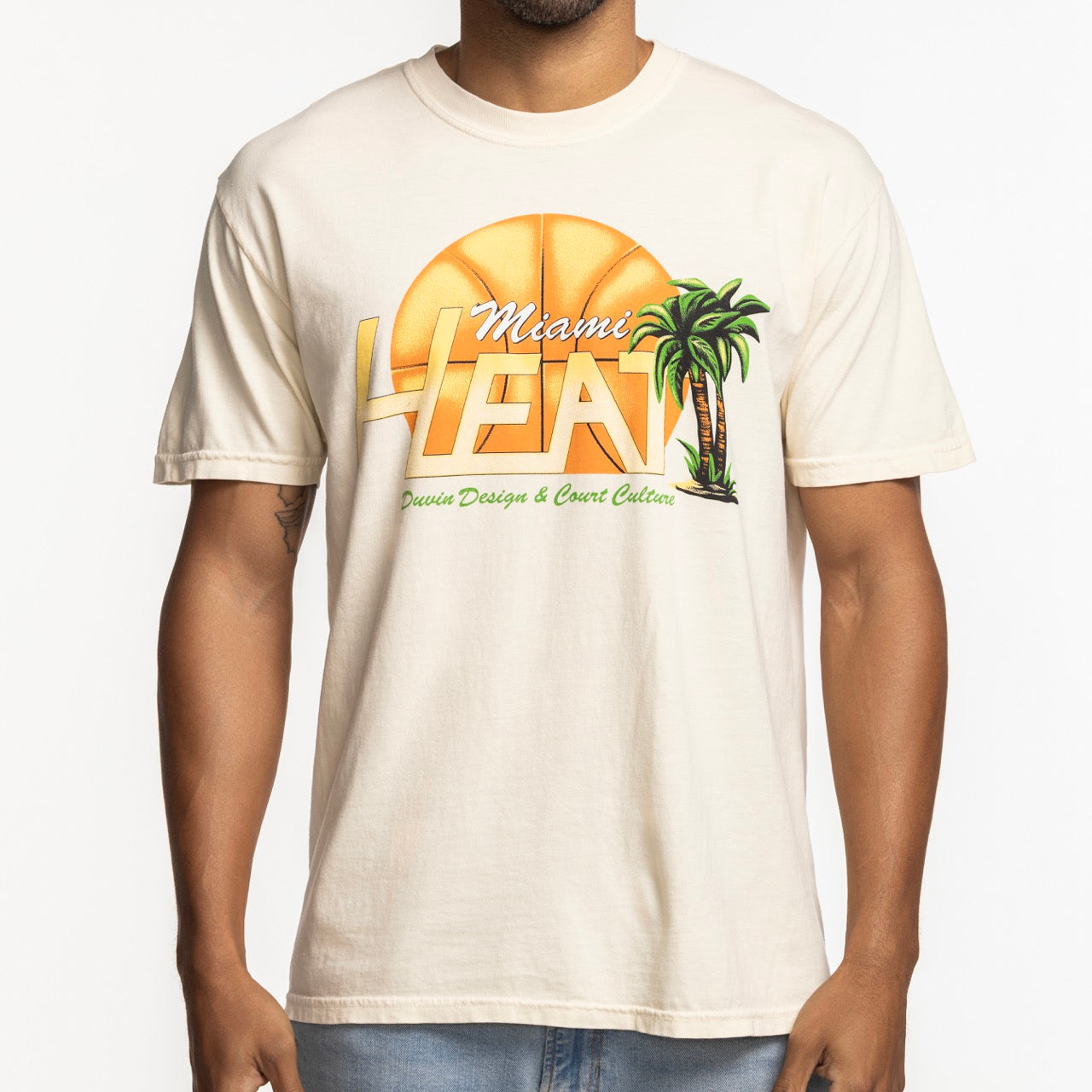 A person wears the Court Culture x Duvin Flames Tee by Court Culture—a cream midweight cotton T-shirt with a large basketball graphic, two palm trees, and bold Miami HEAT lettering from the exclusive collection.