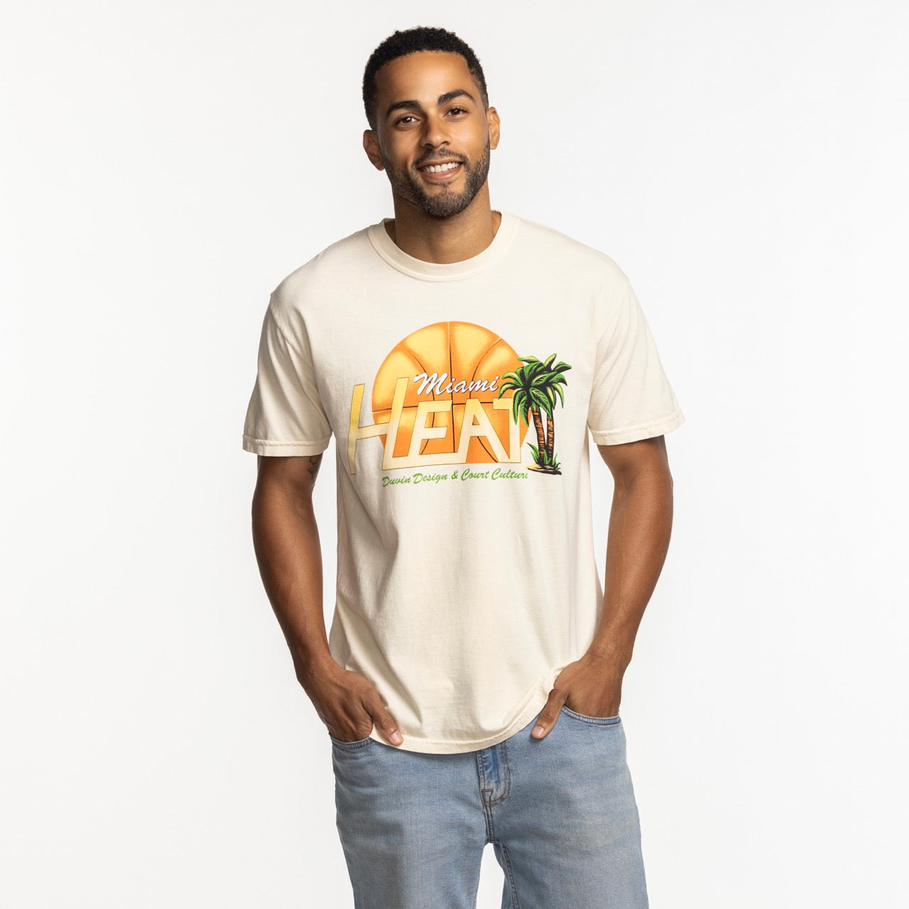 A man smiles in a beige Court Culture x Duvin Flames Tee, featuring a sunset, palm trees, and basketball graphic. The pre-shrunk midweight cotton tee is styled with light blue jeans against a white background.