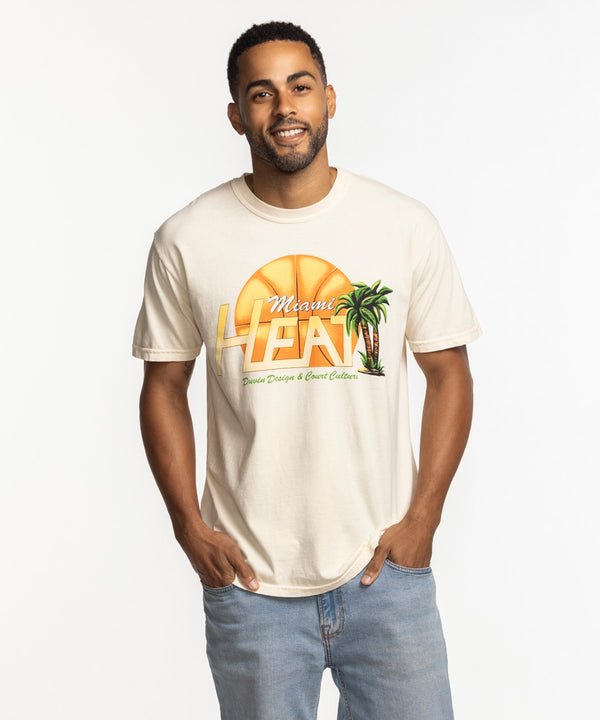 A man smiles in a beige Court Culture x Duvin Flames Tee, featuring a sunset, palm trees, and basketball graphic. The pre-shrunk midweight cotton tee is styled with light blue jeans against a white background.