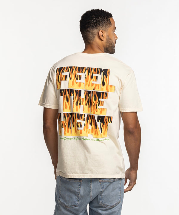 A man smiles in a beige Court Culture x Duvin Flames Tee, featuring a sunset, palm trees, and basketball graphic. The pre-shrunk midweight cotton tee is styled with light blue jeans against a white background.