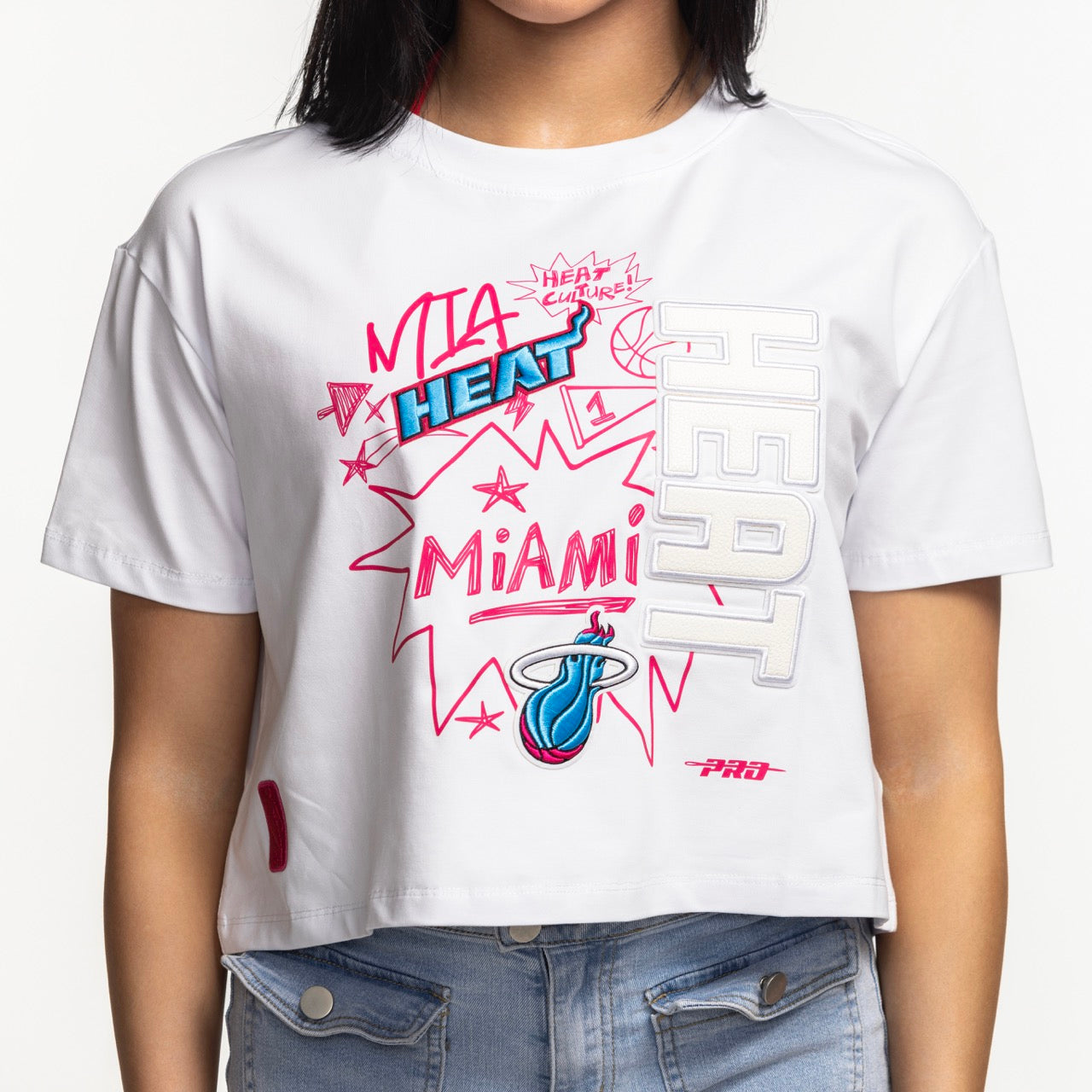 A woman wears the Pro Standard Miami HEAT Original Vice Women's Boxy Tee by Pro Standard, a white cotton spandex crop top with colorful graphic text, star accents, and team logo, paired with light blue jeans. Wash with like colors.