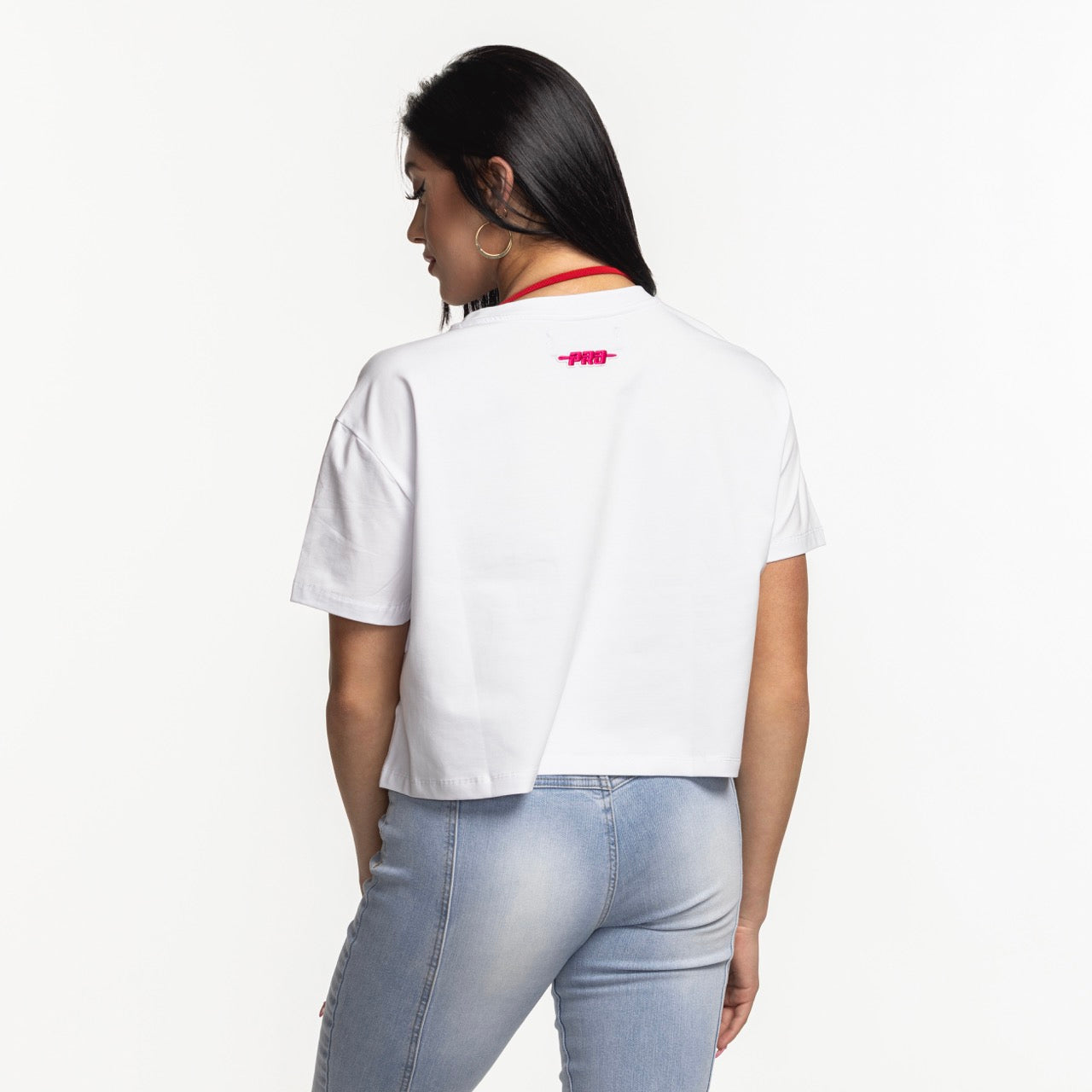 A woman with long dark hair wears the Pro Standard Miami HEAT Original Vice Women's Boxy Tee by Pro Standard, a relaxed fit white cropped t-shirt in soft cotton spandex with a small pink graphic on the back, paired with light blue jeans.