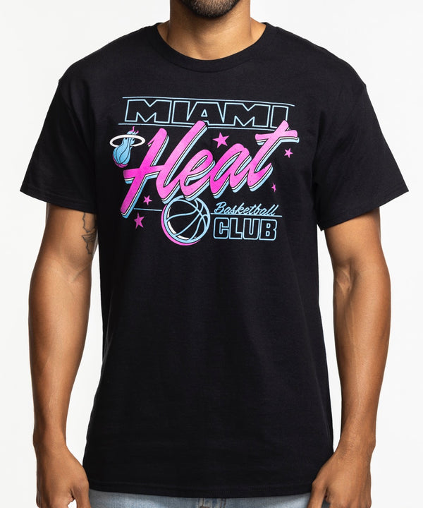 A man with short hair and a beard smiles while wearing the Item Of The Game Miami HEAT Original Vice Nights Basketball Club Tee, featuring pink, blue, and white graphics, and light blue jeans against a plain white background.
