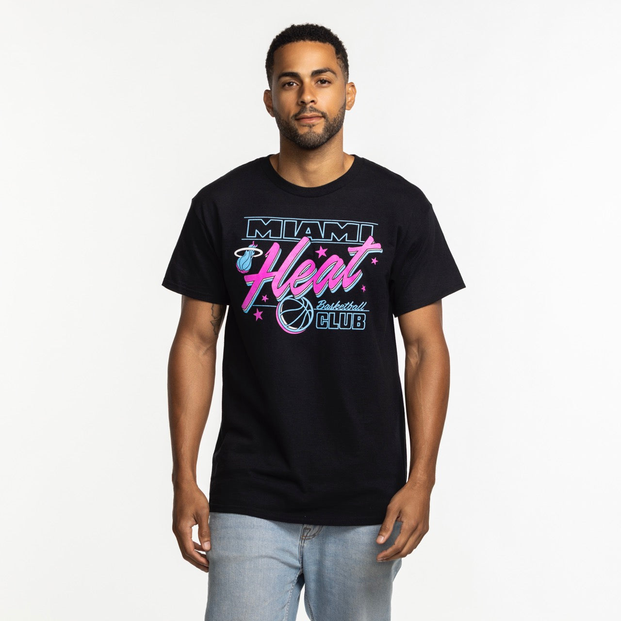A man faces forward wearing the Item Of The Game Miami HEAT Original Vice Nights Basketball Club Tee, featuring neon-style graphics, paired with light blue jeans against a plain white background.