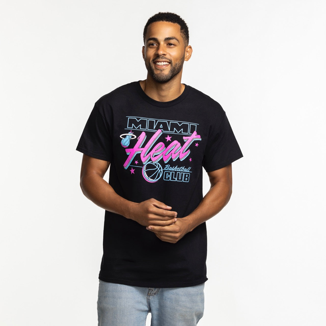 A man with short hair and a beard smiles while wearing the Item Of The Game Miami HEAT Original Vice Nights Basketball Club Tee, featuring pink, blue, and white graphics, and light blue jeans against a plain white background.