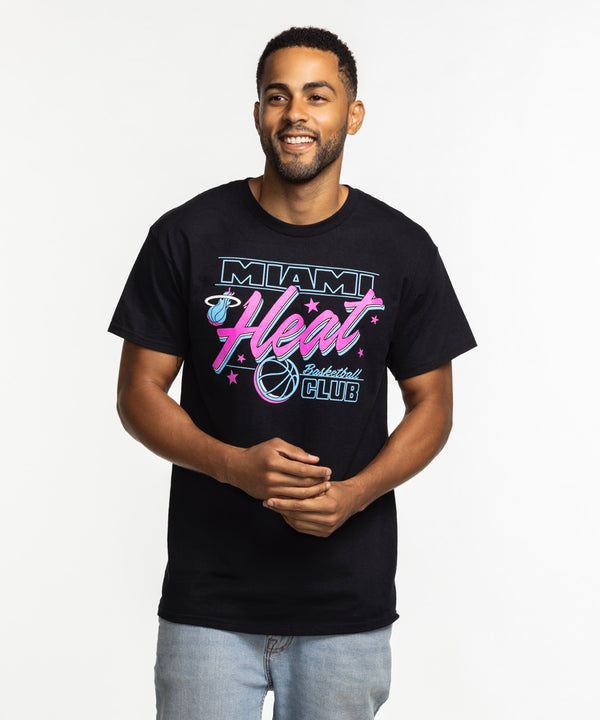 A man with short hair and a beard smiles while wearing the Item Of The Game Miami HEAT Original Vice Nights Basketball Club Tee, featuring pink, blue, and white graphics, and light blue jeans against a plain white background.