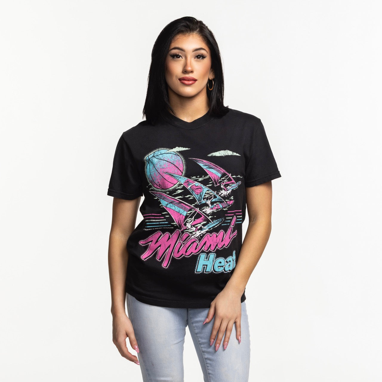 A woman with straight black hair wears the Court Culture x Duvin Tropical Tee by Court Culture—a black graphic t-shirt with neon pink and blue accents—paired with light blue jeans, standing against a plain white background facing forward.