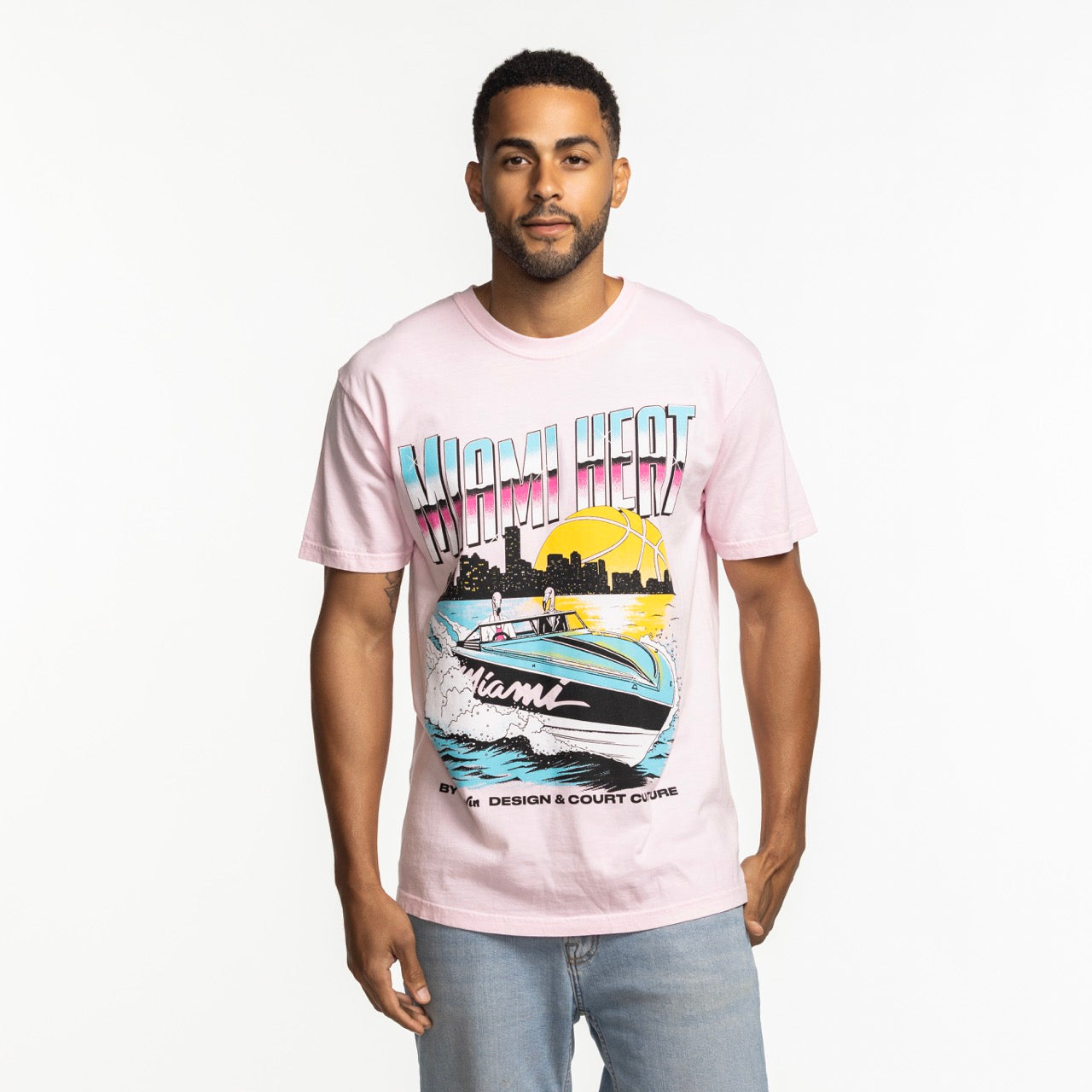 A man stands against a white background wearing the Court Culture x Duvin Boat Tee by Court Culture, a light pink pre-shrunk midweight cotton shirt with a speedboat, sunset, palm trees, and 